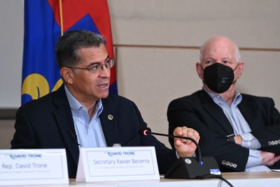 Secretary Becerra is seated at a conference table wearing a dark blazer and blue shirt. To the right of him is a gentleman wearing a dark blazer, blue shirt and black maskm