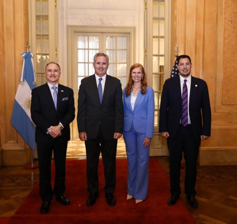 Ambassador Peter Lamelas, Minister Pablo Quirno, Director Bethany Kozma, Secretary’s Office of Global Affairs and Agustín Caulo, Secretary of Worship and Civilization stand side‑by‑side on a red carpet in a room with flags of Argentina and the United States displayed behind them.