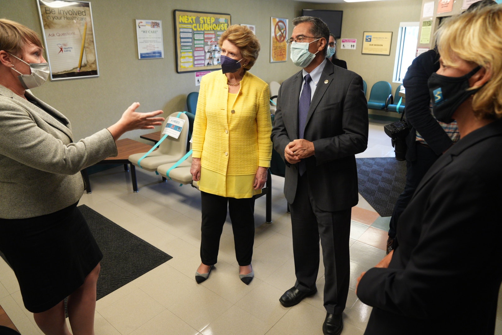 Secretary Becerra is wearing a dark suit with dark rimmed reading glasses and mask, to the right of him is a women wearing a yellow blouse with a dark color mask, in front of Secretary Becerra is a lady wearing a neutral color suit and mask