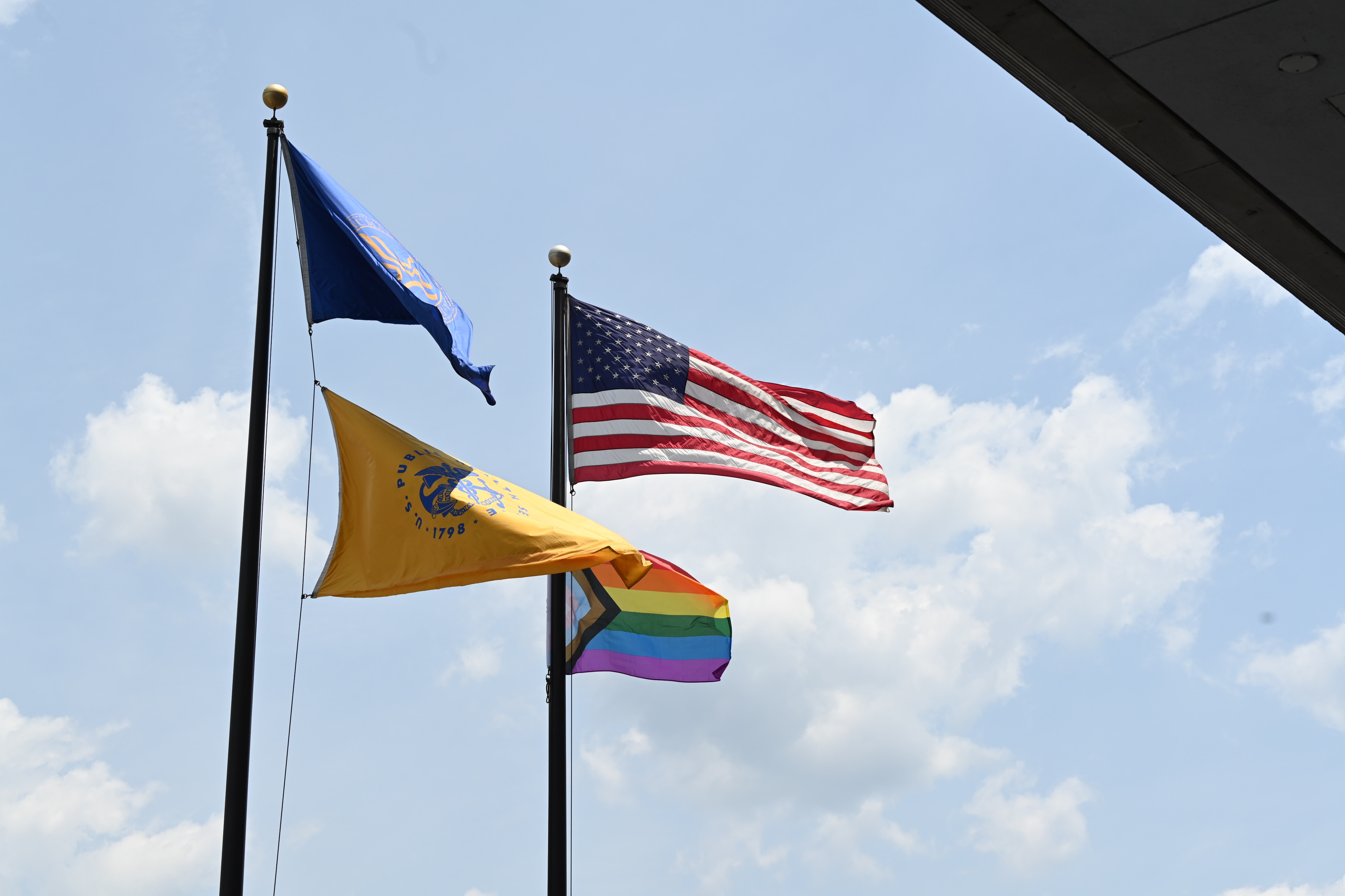 Progress Pride flag, American flag, HHS flag and Public Health Service flag waving in front of a blue sky with large white clouds.