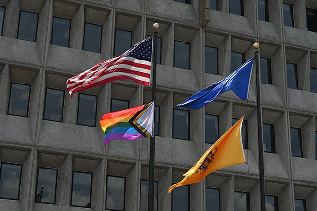 Close up photo of the Progress Pride flag, American flag, HHS flag and Public Health Service flag waving in the wind outside of HHS headquarters building on June 1, 2022.
