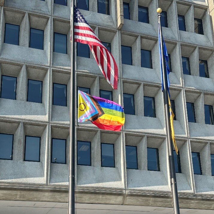 Close up photo of the Progress Pride flag and American flag waving in the wind outside of HHS headquarters building.