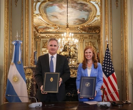 Minister Pablo Quirno and Director Bethany Kozma Secretary’s Office of Global Affairs stand side‑by‑side in a room with gold detailing, holding up signed documents for a photo.