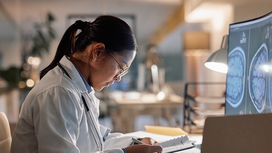 A person wearing a white lab coat sits at a desk reviewing notes while looking at brain scan images displayed on a computer monitor in a clinical or research setting.
