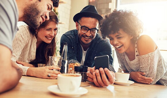 A group of young adults gathered around a kitchen table looking at a mobile phone.