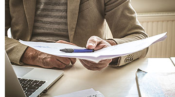 Image of person at a desk with a laptop holding a piece of paper and a pen.