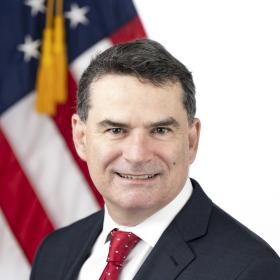Sean McCluskie is seated in front of the American flag wearing a dark colored suite with a dark red tie.