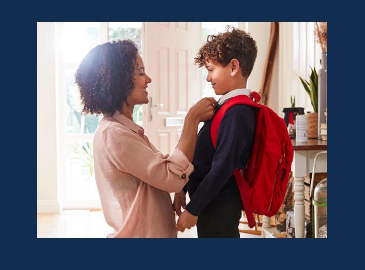 Adult helping child put on a red backpack in the entryway of home.